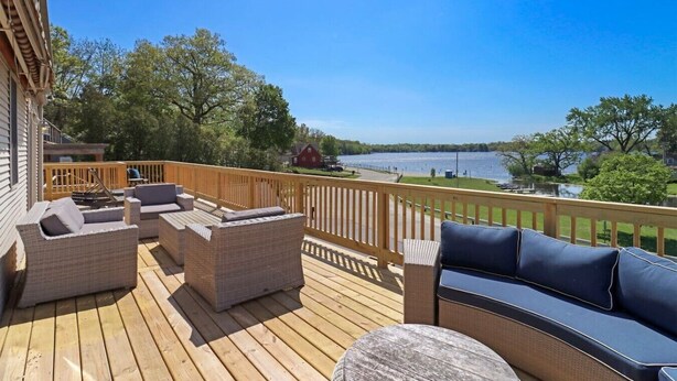 Une terrasse en bois avec des meubles d’extérieur, une vue sur un lac et un bâtiment rouge en arrière-plan.