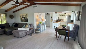 Un vaste espace de vie doté d’un plafond haut, de poutres en bois, d’un ensemble de canapés, d’une table à manger avec des chaises, et d’une cuisine en arrière-plan.