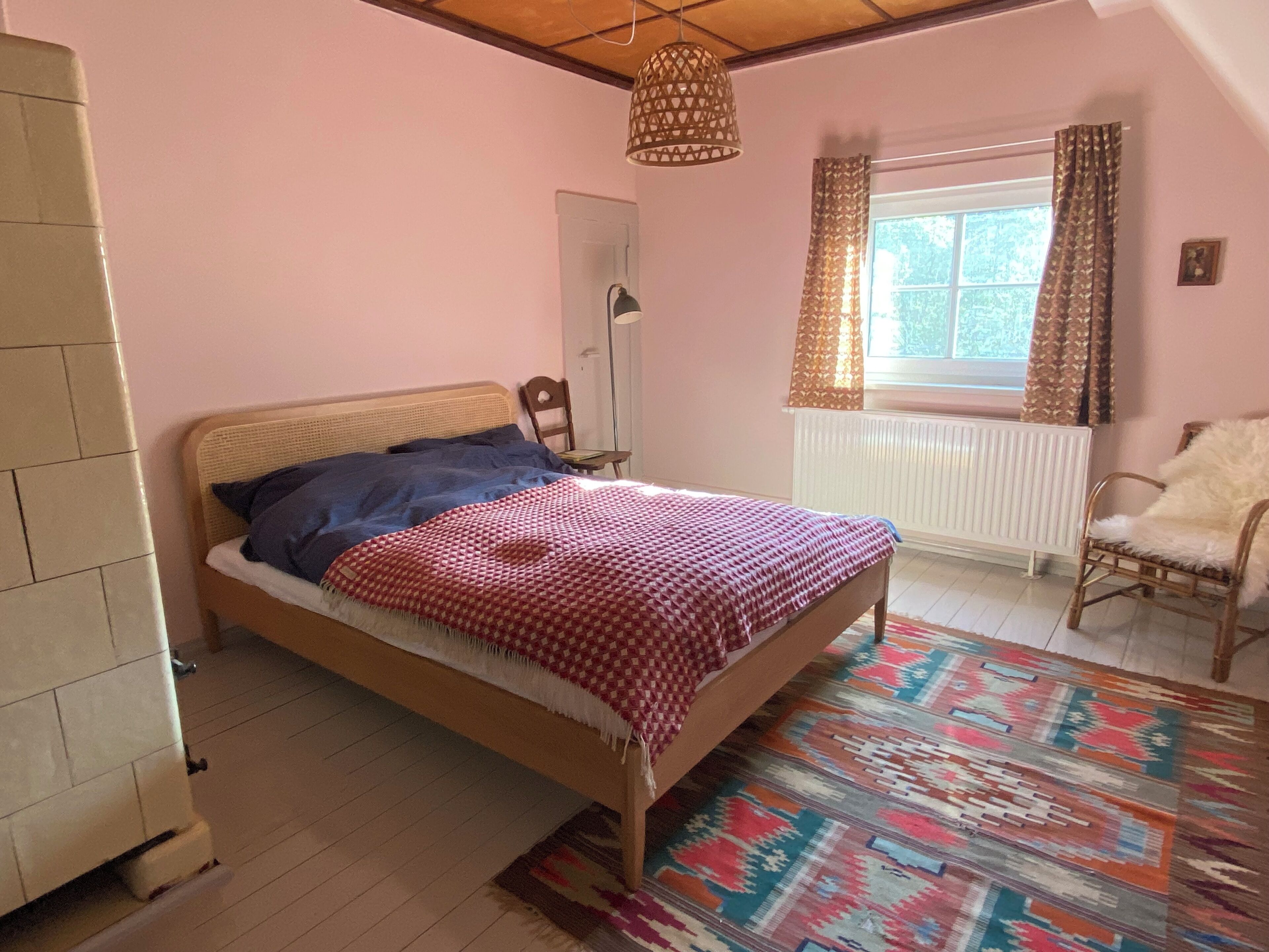 Une chambre à coucher comprenant un lit, une chaise, un radiateur, une fenêtre avec des rideaux et un luminaire suspendu.
