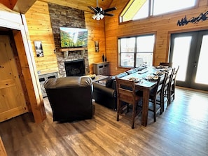 A log cabin interior with a fireplace, television, dining area, and a view of the outdoors. - Views from Deck-Hot Tub, game room, 5 min to parkway and attractions. (Pigeon Forge)