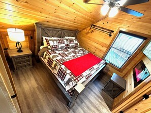 Quarto com cama, mesa de cabeceira, abajur, ventilador de teto e janela com persianas.
