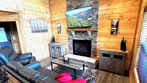 A log cabin interior with a fireplace, a television mounted above it, a leather sofa, and a small table with decorative items. - Views from Deck-Hot Tub, game room, 5 min to parkway and attractions. (Pigeon Forge)