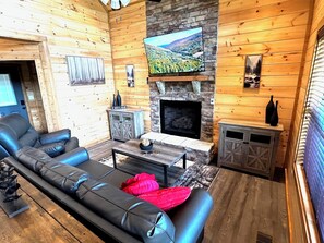 A log cabin interior with a fireplace, a television mounted above it, a leather sofa, and a small table with decorative items. - Views from Deck-Hot Tub, game room, 5 min to parkway and attractions. (Pigeon Forge)
