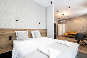 Una habitación de hotel moderna con una cama grande, cabecera de madera, pared con un diseño geométrico, una zona de comedor con un sofá amarillo y una pequeña mesa con una silla.