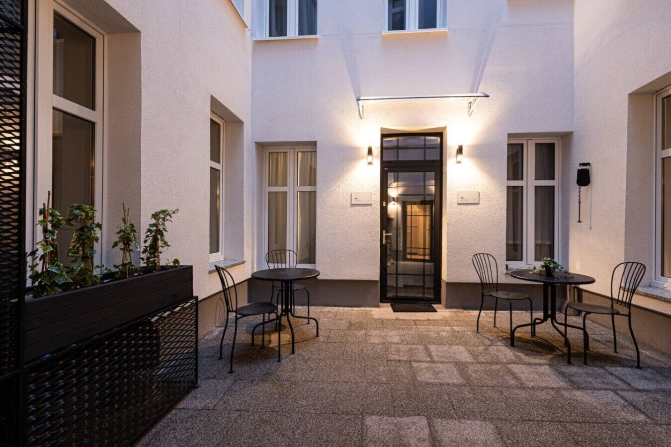 Une cour avec une porte, deux tables accompagnées de chaises et un jardinier avec des plantes.