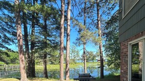 A patio with a grill, chairs, and a view of a lake surrounded by tall trees.