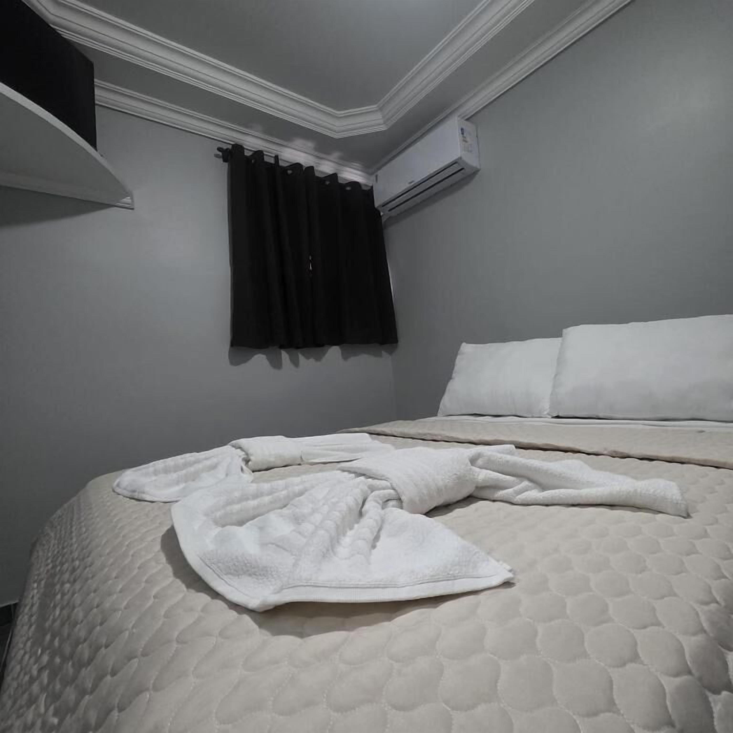 A bedroom with a bed, pillows, a blanket, a dark curtain, and an air conditioning unit.