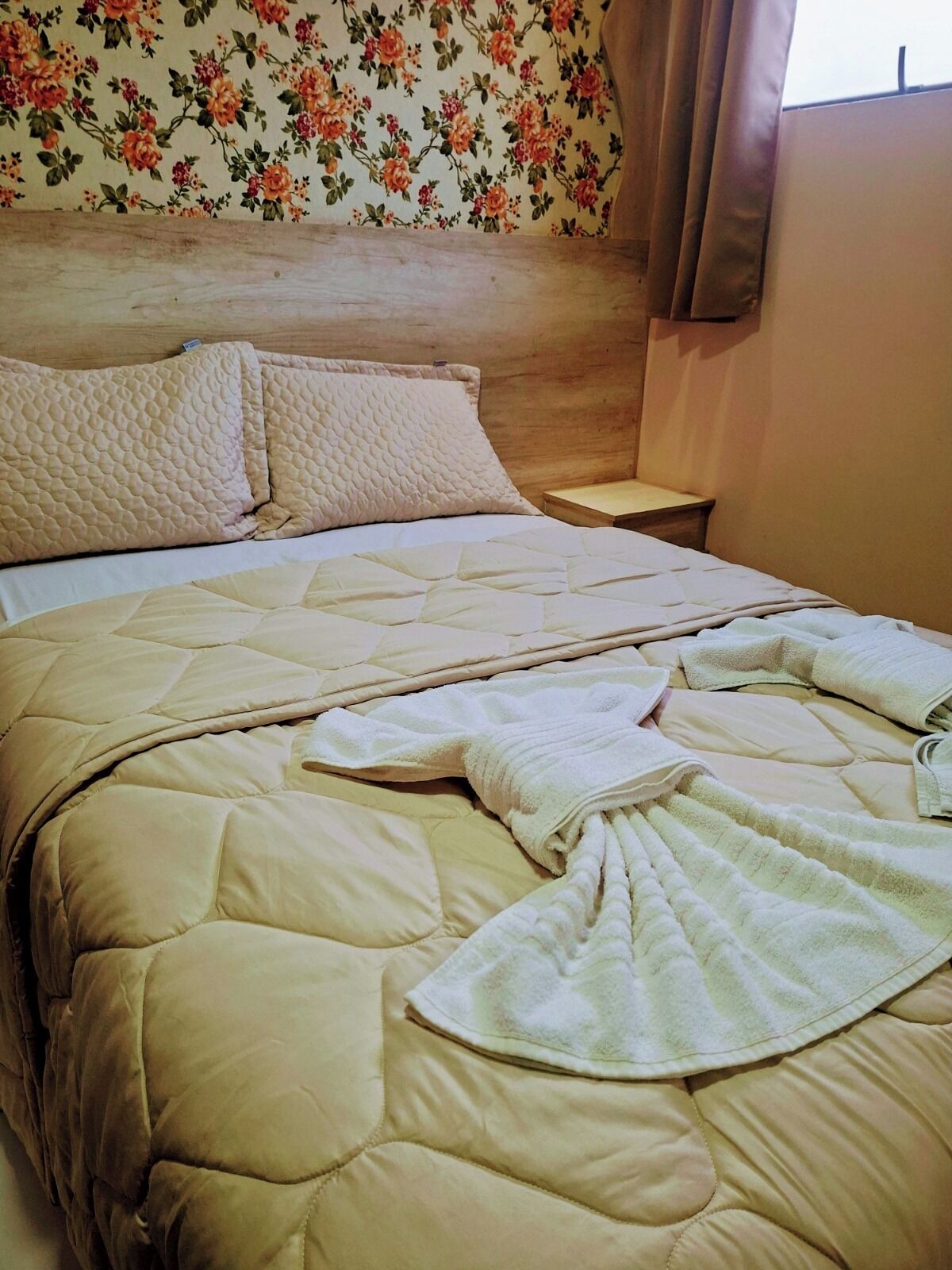 A neatly made bed with a quilted headboard, a bedside table, and a floral wallpaper pattern.
