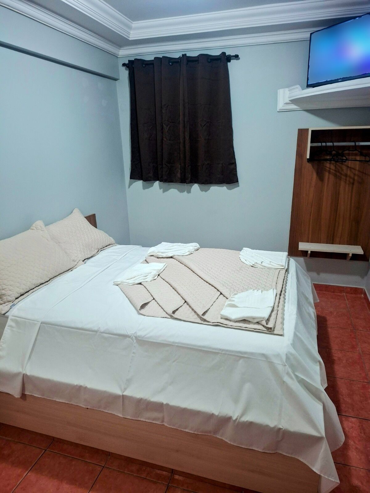 A neatly made bed with a white bedspread and pillows, a black curtain, and a television mounted on the wall.