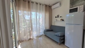 Ein Wohnzimmer mit einer Couch, einem Kühlschrank und einem Fenster mit durchsichtigen Vorhängen.