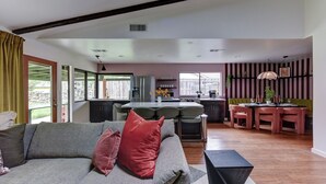 A modern living room with a sofa, a dining area with chairs, a kitchen with a bar, and large windows.