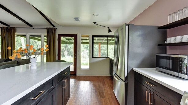 A modern kitchen with dark cabinetry, a stainless steel refrigerator, a microwave, and a large window with curtains.