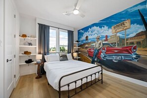 A bedroom with a bed, a ceiling fan, a window with curtains, a shelf with hats, and a mural of a vintage car and a man. - Suite Duplex @Downtown Unit B 6 Br/6 Bath (Houston)