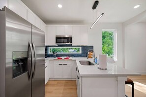 A modern kitchen with white cabinetry, stainless steel appliances, a built-in water dispenser, and a large window providing a view of greenery. - Suite Duplex @Downtown Unit B 6 Br/6 Bath (Houston)