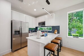 A modern kitchen with white cabinetry, stainless steel appliances, a central island with stools, and a window with a view of greenery. - Suite Duplex @Downtown Unit B 6 Br/6 Bath (Houston)