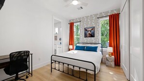 Una camera da letto con un letto, una scrivania, una sedia, una finestra con tende e una ventola a soffitto.
