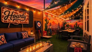 Una zona relax all'aperto accogliente, con un divano blu, un tavolo con piano in vetro e un elemento decorativo a fiamma, e un murale raffigurante cactus e paesaggi desertici.