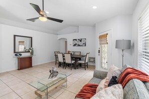 A living room with a glass coffee table, a sofa with a red blanket, a dining area with chairs and a table, and a ceiling fan.