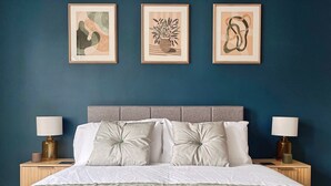 Ein ordentlich bezogenes Bett mit einem gemusterten Überwurf, zwei Nachttischlampen und drei gerahmten Kunstwerken an der Wand.