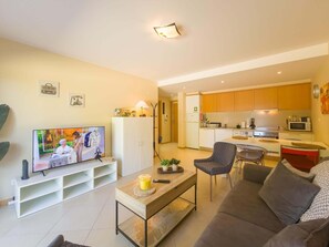 Ein modernes Wohnzimmer mit Flachbildfernseher, einer Couch, einem Couchtisch und einer Küche im Hintergrund.