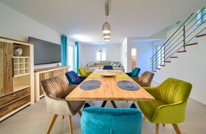 Ein modernes Wohnzimmer mit einem Holzesstisch, bunten Stühlen, einem Sofa und einem Fernseher.