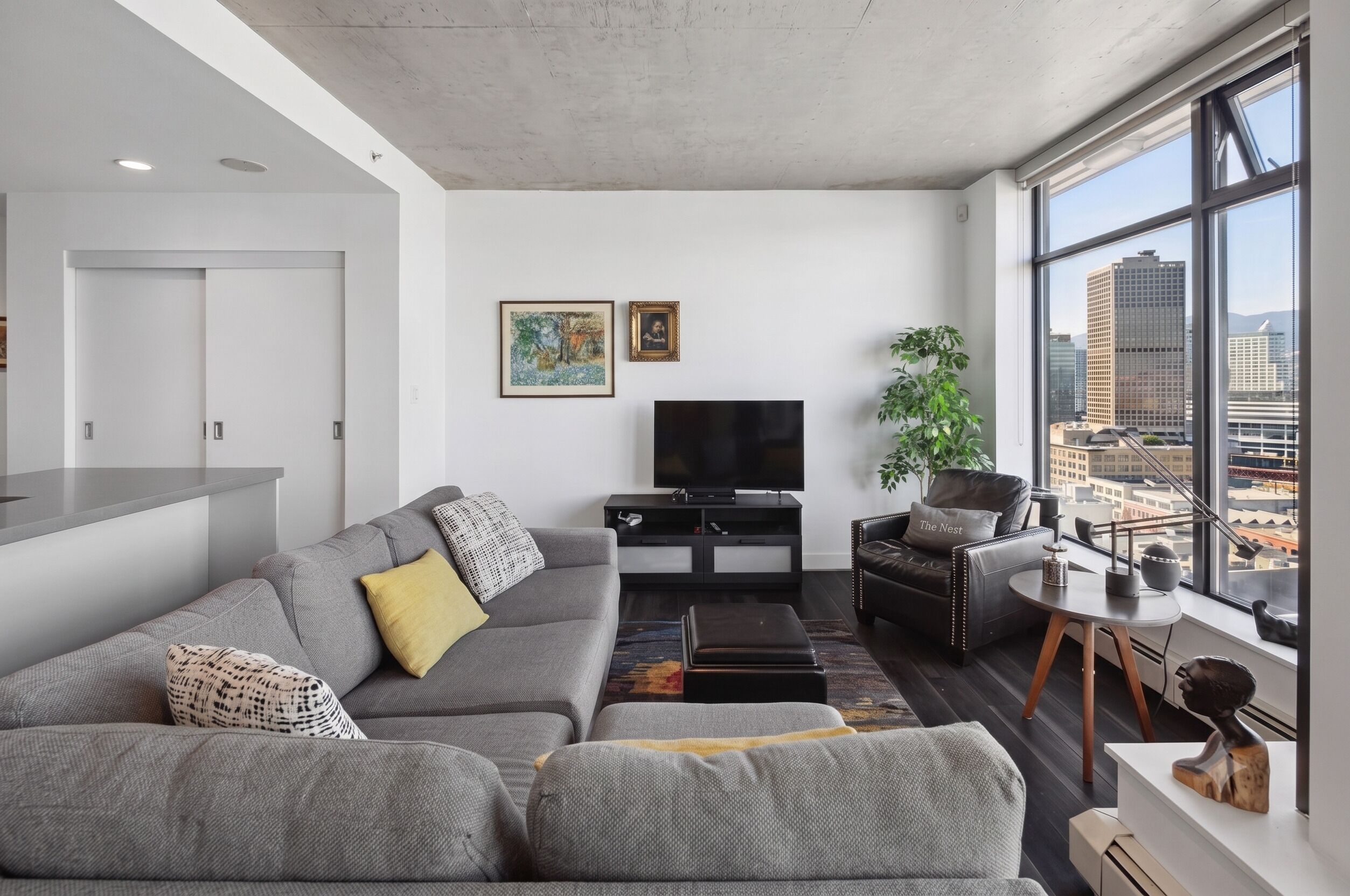 Un salon moderne avec un canapé gris, un meuble de rangement noir pour les appareils électroniques et une grande fenêtre donnant sur la ville.