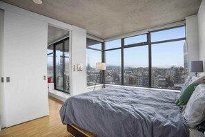 A modern bedroom with a large bed, a view of the city through floor-to-ceiling windows, and a sliding door leading to a balcony.