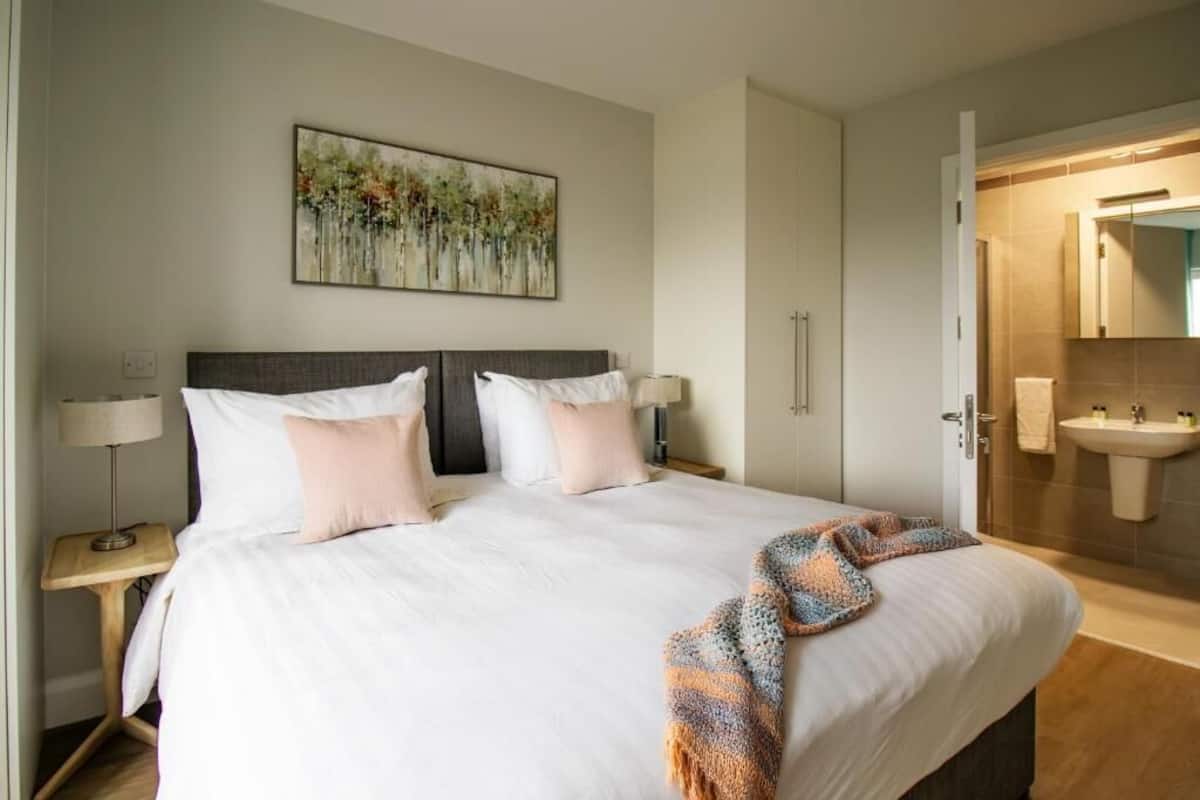 Habitación de hotel con una cama grande, una mesita de noche con lámpara, un baño con lavamanos y espejo, y un cuadro en la pared.