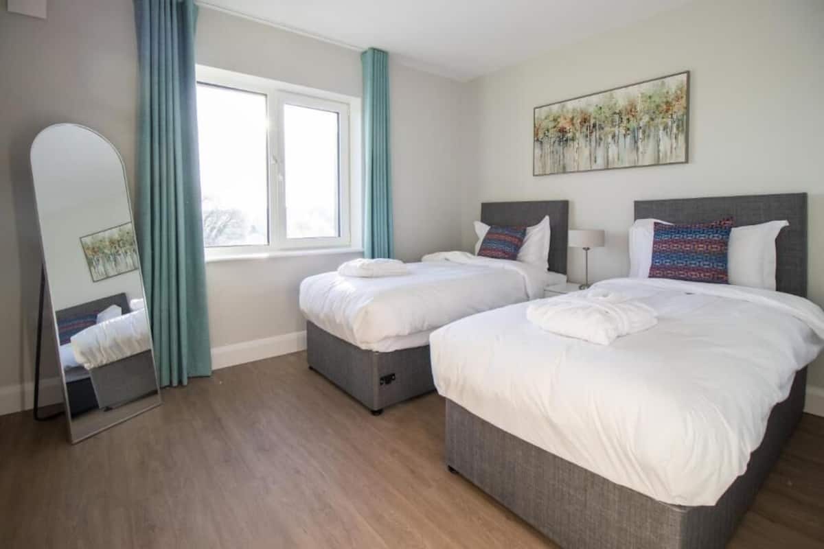 Habitación de hotel con dos camas, un ventanal con cortinas, un espejo y un cuadro en la pared.