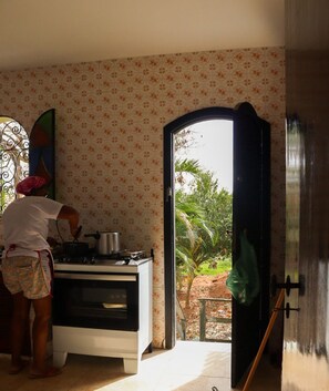 Eine Küche mit Herd, Topf und einer Person beim Kochen. Ein Fenster bietet Blick auf Grünflächen.