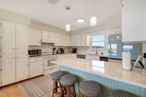 A modern kitchen with white cabinetry, a large island, bar stools, and a view of the sea through a window.