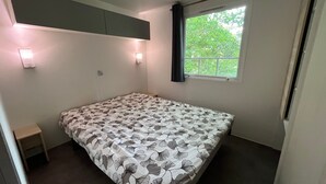 Ein kleines Zimmer mit einem Einzelbett, einer Bettdecke mit Muster, einem Fenster mit Vorhängen und einem Schrank über dem Bett.