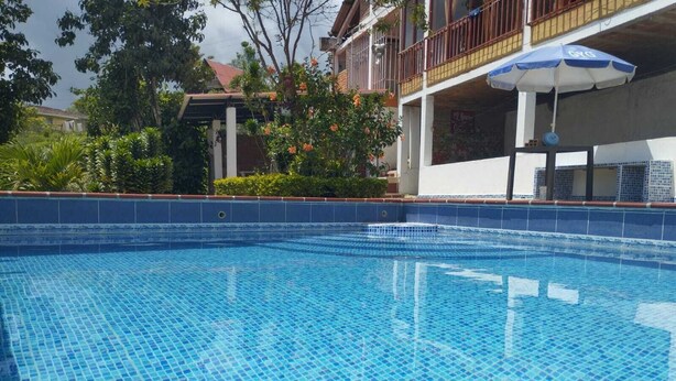 A clear blue pool with a tiled edge, surrounded by greenery and a building with a balcony.