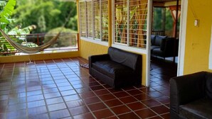 A room with a tiled floor, a black sofa, a hammock, and a view of greenery through a window with metal bars.