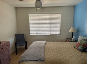 Ein Schlafzimmer mit einem Bett, einem Sessel, einer Kommode, einem Fenster mit Jalousien, einer Lampe und einem Deckenventilator.