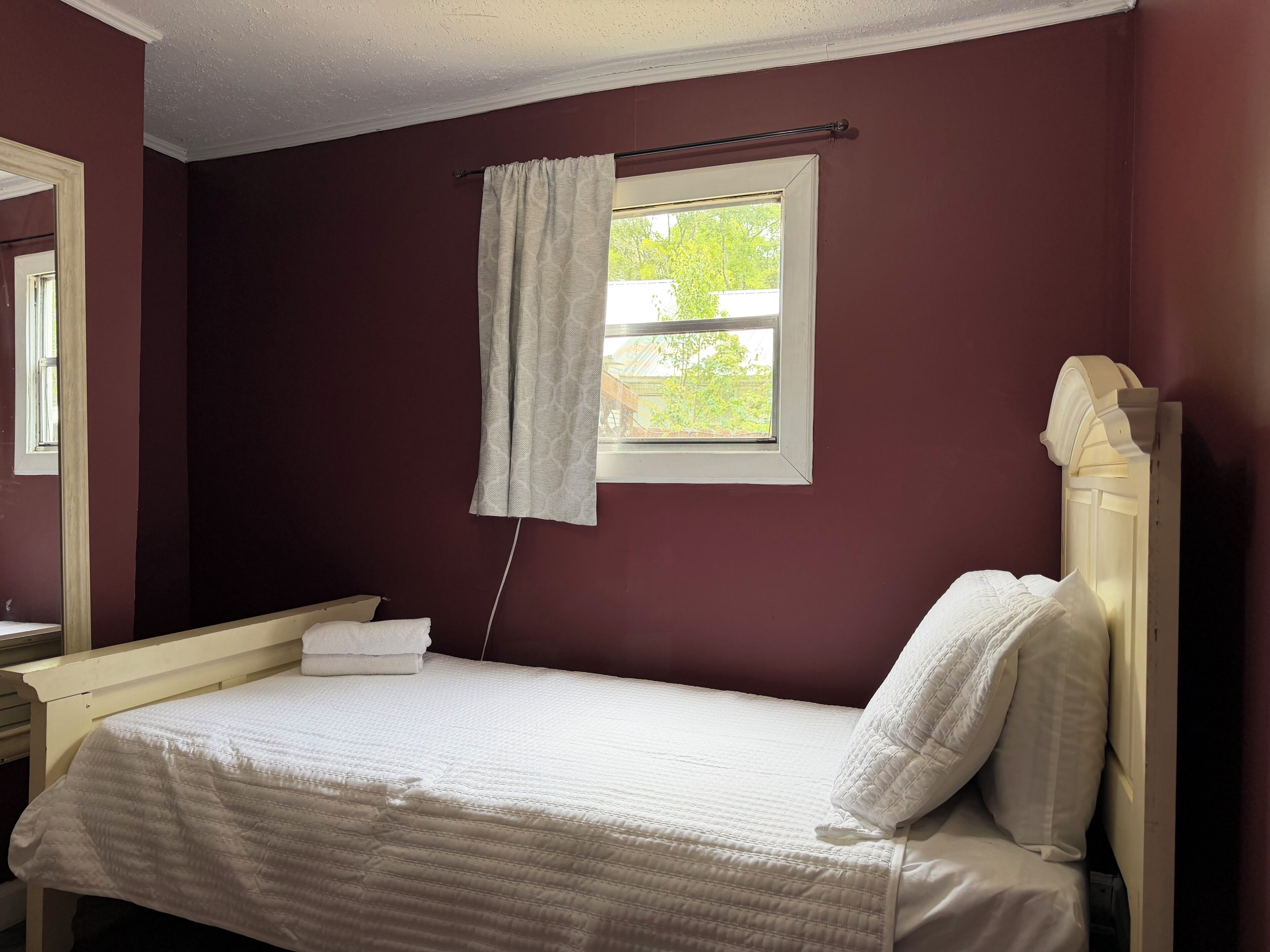 A bedroom with a single bed, a bedside table, a window with curtains, and a mirror.