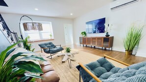 Un salon moderne avec un canapé en cuir marron, un fauteuil gris et une table basse en bois. Une grande fenêtre laisse entrer la lumière naturelle, un téléviseur à écran plat est fixé au mur et une plante en pot se trouve sur le côté droit.
