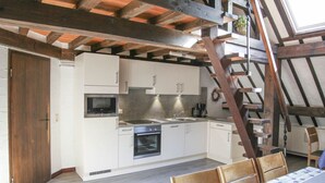 Una cocina con armarios blancos, microondas, horno y lavamanos. Se ve una escalera de madera apoyada contra la pared, y una mesa con sillas al frente.