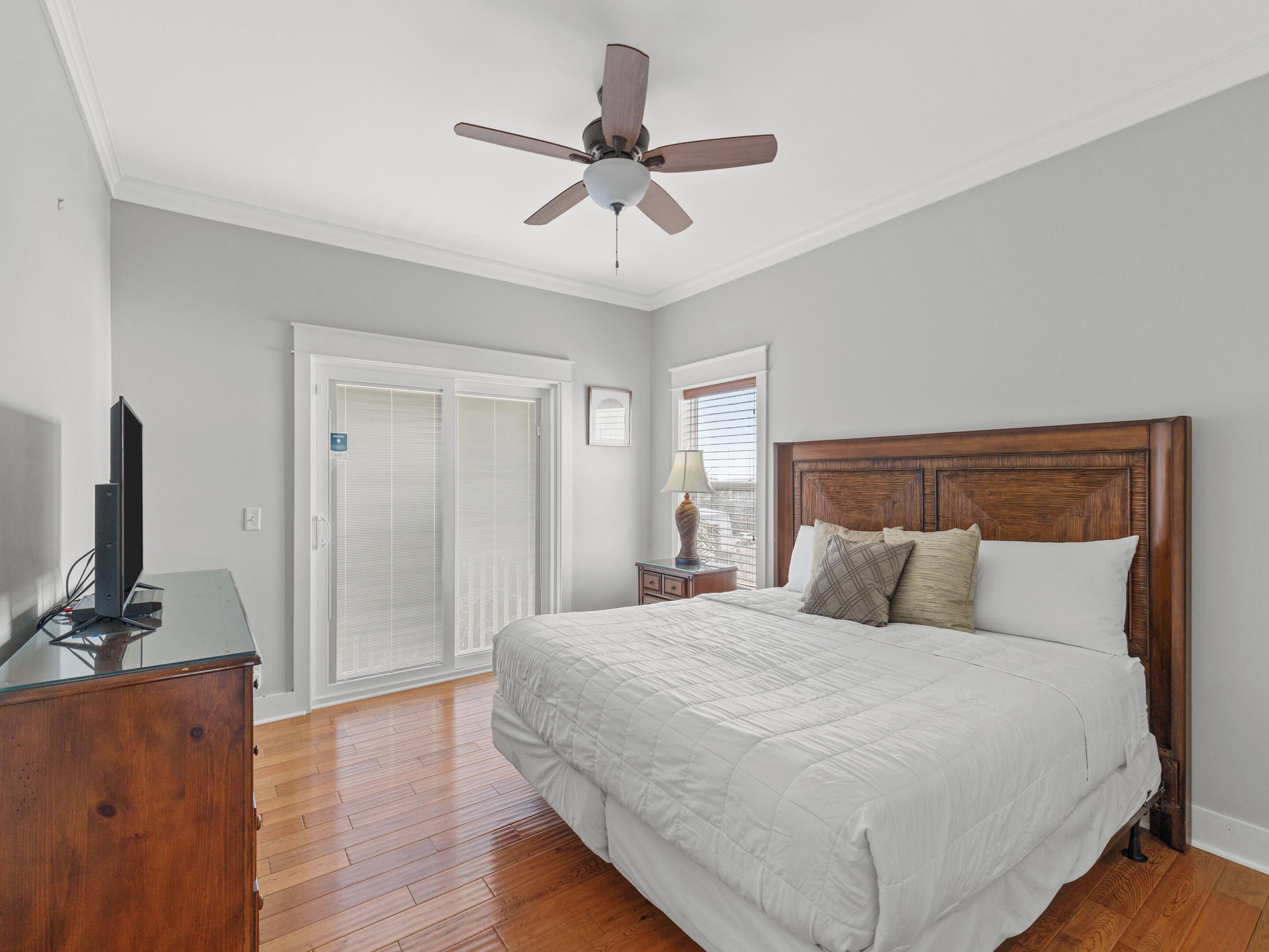 A bedroom with a bed, a ceiling fan, a TV, a wooden dresser, and a sliding glass door.