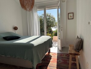 A bedroom with a bed, a hanging light fixture, a door leading to a balcony, and a small wooden stool. - Rental mini villa 180° sea view Palombaggia Porto Vecchio (Porto-Vecchio)