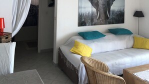 Un dormitorio moderno con una cama blanca, almohadas turquesa y amarillas, una lámpara colgante de madera, un cuadro enmarcado con un elefante y una silla de mimbre.