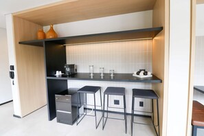Uma cozinha compacta com bancada preta, três banquetas, uma geladeira pequena e dois vasos laranjas em uma estante.