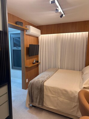 Une chambre à coucher avec un lit, un téléviseur fixé au mur, une table de chevet avec un téléphone et une fenêtre avec des rideaux.