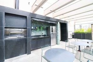 Une cuisine extérieure équipée d’un barbecue encastré, d’un réfrigérateur et d’un évier. On y trouve également des tables et des chaises d’extérieur.