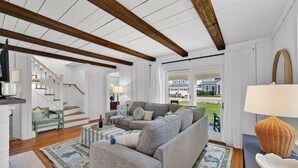 A spacious living area with a large grey sofa, wooden beams on the ceiling, a staircase, and a view of a house through a glass door. - Steps To Peggotty Beach! Slps 10 + Dogs OK! (Scituate)
