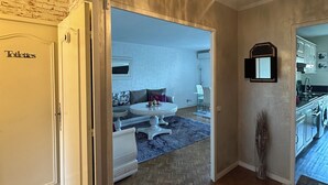 Ein Korridor mit Fischgrätparkett, einer Chandelieber, einer Tür mit der Aufschrift 'Toiletten' und einem Blick in ein Wohnzimmer mit Sofa und Tisch.
