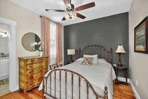 A bedroom with a wooden bed, a dresser, a ceiling fan, a mirror, and a window with curtains.