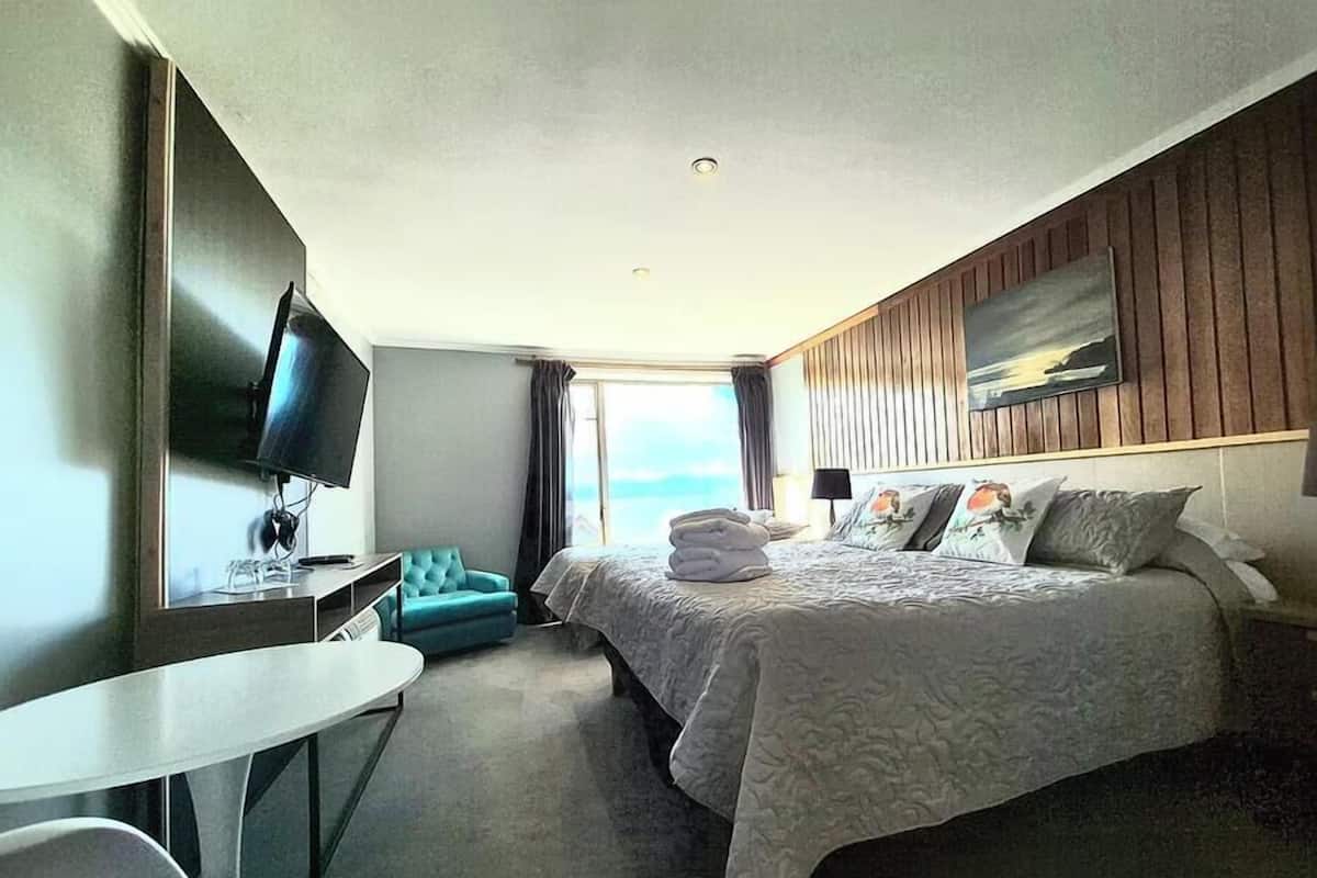 A hotel room with a bed, a desk, a TV, and a view of the ocean.