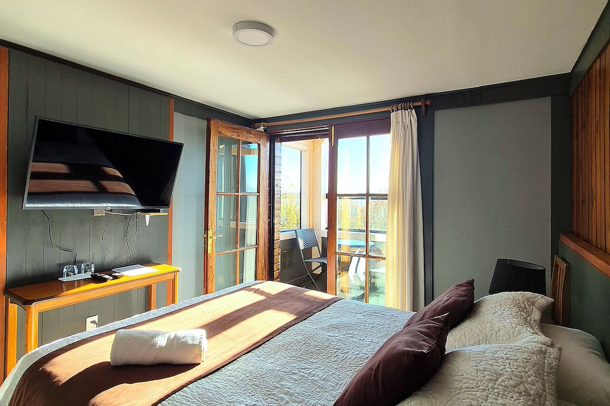 A hotel room with a bed, a TV, a desk with a chair, and a balcony with a view.
