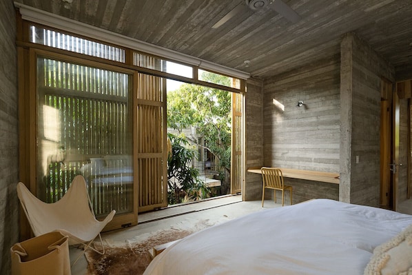 A bedroom with a bed, a chair, a desk, and a sliding glass door leading to a garden. - Casa Macu Saladita Surf House (Ixtapa)